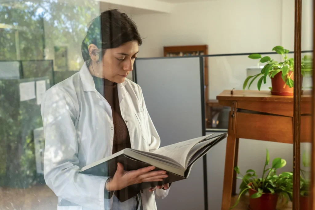 Woman in a white coat reading a book beside a window
